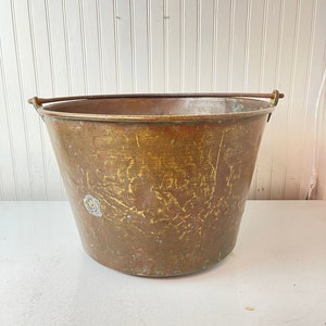 May include: A vintage brass bucket with a copper handle. The bucket has a worn, aged patina and is perfect for a rustic or farmhouse decor.
