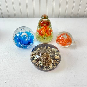 May include: Four glass paperweights with different designs. One is a clear glass sphere with a blue swirl inside. Another is a clear glass sphere with a brown pine cone inside. The third is a clear glass sphere with an orange flower inside. The fourth is a clear glass cone with an orange flower inside.
