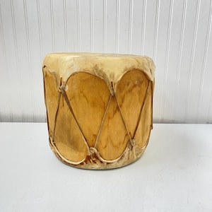 May include: A brown leather-covered drum with a wooden frame and a laced design. The drum is sitting on a white surface.