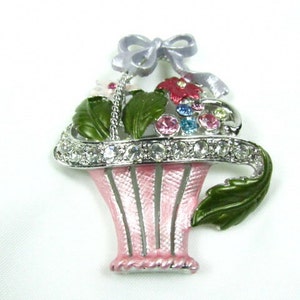 May include: A silver tone brooch with a pink and white enamel basket filled with colorful flowers and rhinestones. A silver tone bow with a lavender ribbon is attached to the top of the basket.