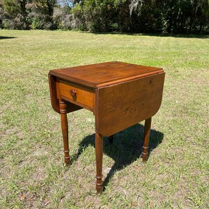 May include: A small, antique wooden drop-leaf table with a single drawer and turned legs. The table is a warm brown color and is set outdoors on a grassy lawn. The table's leaves are down, and the drawer has a round wooden knob.