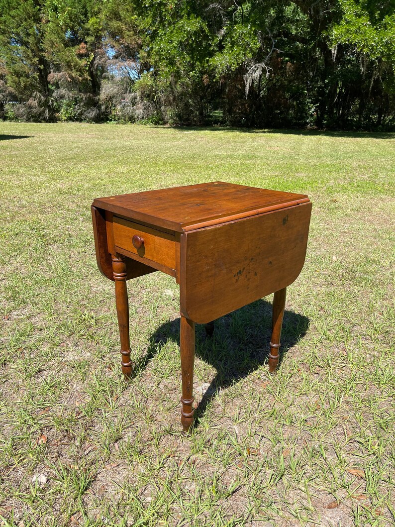 May include: A small, antique wooden drop-leaf table with a single drawer and turned legs. The table is a warm brown color and is set outdoors on a grassy lawn. The table's leaves are down, and the drawer has a round wooden knob.
