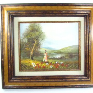 May include: A framed oil painting depicting a woman in a pink dress standing in a field of red and white flowers near a lake. The background features a green hillside and a blue sky.