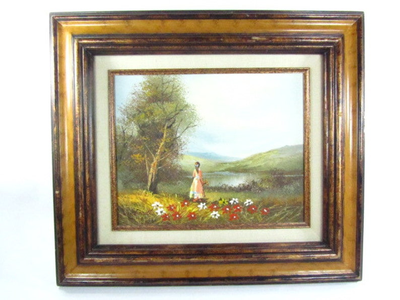 May include: A framed oil painting depicting a woman in a pink dress standing in a field of red and white flowers near a lake. The background features a green hillside and a blue sky.