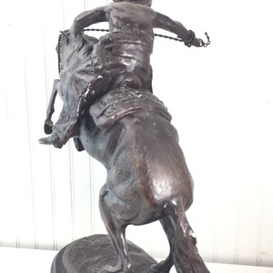 May include: Bronze sculpture of a cowboy riding a bucking horse. The cowboy is wearing a hat and is holding a rope. The horse is rearing up on its hind legs. The sculpture is on a black base.