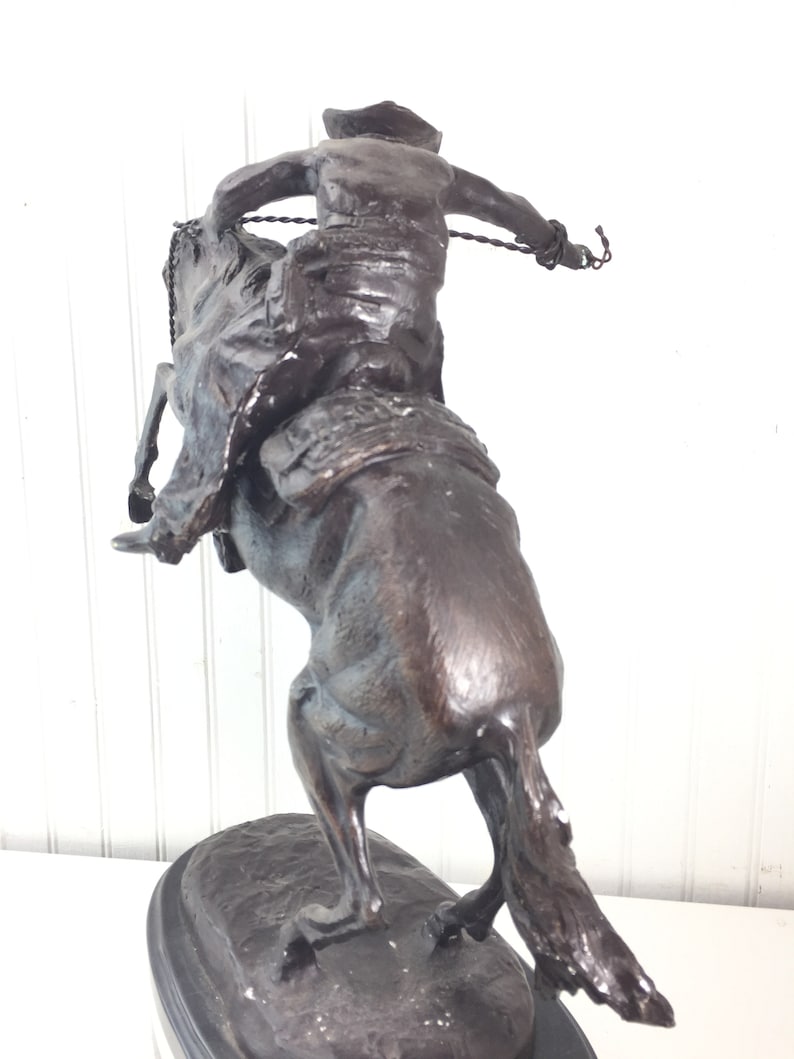 May include: Bronze sculpture of a cowboy riding a bucking horse. The cowboy is wearing a hat and is holding a rope. The horse is rearing up on its hind legs. The sculpture is on a black base.