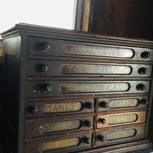 May include: Antique wooden cabinet with eight drawers, each with a glass label and black knobs. The labels are handwritten with names like "James" and "Arthur".