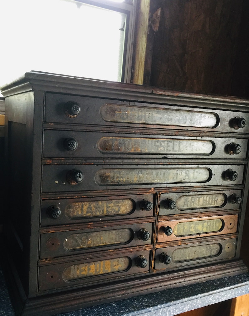 May include: Antique wooden cabinet with eight drawers, each with a glass label and black knobs. The labels are handwritten with names like "James" and "Arthur".