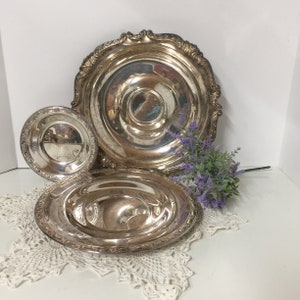 Puede incluir: Tres platos de servir de plata de diferentes tama&ntilde;os, con bordes ornamentados, est&aacute;n dispuestos sobre un encaje de ganchillo blanco. El plato m&aacute;s grande tiene un centro elevado y es el m&aacute;s ornamentado. El plato m&aacute;s peque&ntilde;o es liso y se coloca encima del plato del medio.