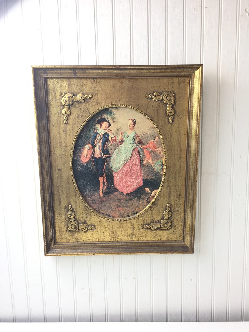 May include: A framed oval print of a couple in 18th century clothing, set against a gold background with ornate corners. The man is wearing a blue coat and hat, and the woman is wearing a pink and green dress.