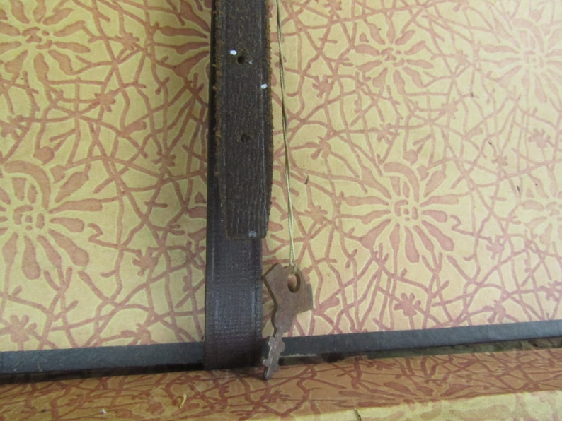 May include: A close-up of a brown leather strap with a metal buckle. The strap is attached to a surface with a patterned design.