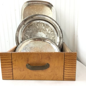 May include: A wooden drawer with a brass handle, containing a stack of three silver serving trays with ornate designs.