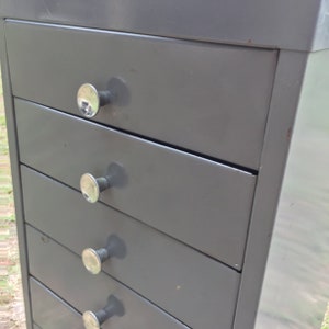 May include: A gray metal cabinet with five drawers and silver metal knobs. The cabinet is made of metal and has a simple design.