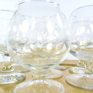 May include: Set of five clear glass brandy snifters with a round bowl and a short stem. The glasses are arranged on a gold-colored surface.