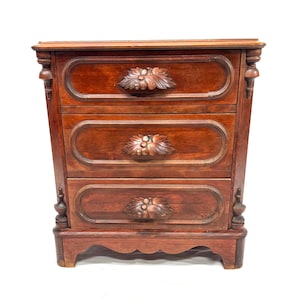 May include: A three-drawer wooden nightstand with a rich, reddish-brown finish. Each drawer features a carved floral design and ornate pulls. The nightstand has decorative side accents and a scalloped base.