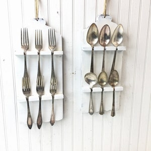 May include: Two white wooden wall-mounted utensil holders with a distressed finish. One holder has four silver forks hanging from it, and the other holder has three silver spoons hanging from it.