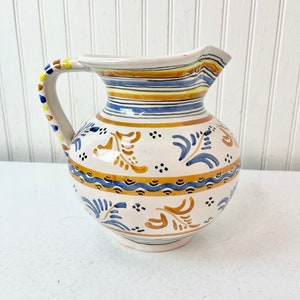 May include: A white ceramic pitcher with a blue and yellow striped design and floral patterns. The pitcher has a handle with a yellow and brown stripe.