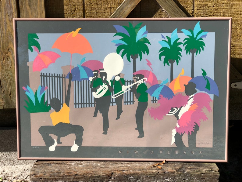 Puede incluir: Una impresi&oacute;n colorida y abstracta de una escena callejera de Nueva Orleans con una banda de m&uacute;sica, palmeras y personas que sostienen paraguas. La impresi&oacute;n est&aacute; enmarcada en un marco rosa claro.