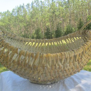 May include: A large, woven basket made of natural materials. The basket is light brown in color and has a rounded, oval shape. It has two handles on either side and is sitting on a white surface.