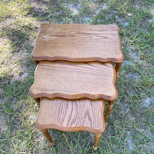 May include: A set of three nesting tables with a light brown wood grain finish. Each table has a scalloped edge and slender legs. The tables are arranged in a stacked configuration, with the smallest table at the bottom.