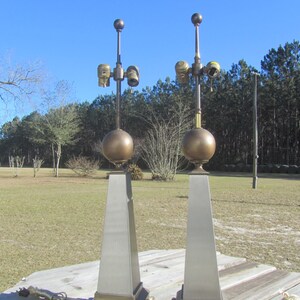 May include: Two tall, silver and gold table lamps with round, gold finials. The lamps have a modern, geometric design and are set on a wooden surface.