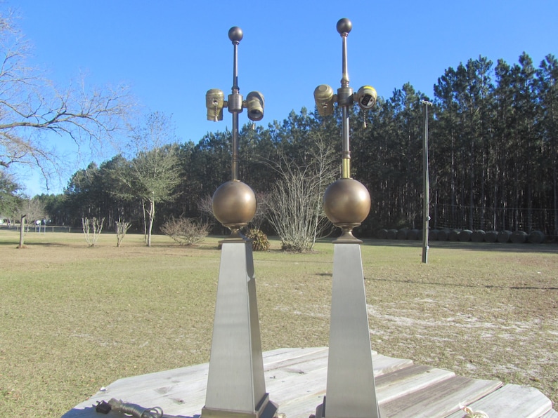 May include: Two tall, silver and gold table lamps with round, gold finials. The lamps have a modern, geometric design and are set on a wooden surface.