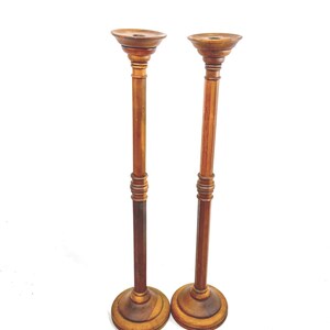 May include: Two tall, wooden candle holders with a classic design. Each has a round base, a long, slender stem, and a small, circular top. The wood has a warm, brown tone, and the pieces appear to be vintage or antique.
