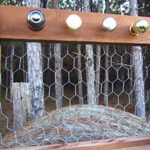 May include: A wooden frame with five different door knobs, including black, silver, and gold finishes. The frame is mounted on a wall with a wire mesh background.