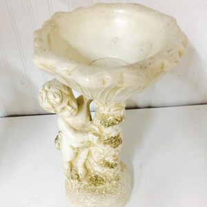 May include: Off-white cherub bird bath. The bird bath features a cherub figure holding up a scalloped bowl. The base and the tree trunk are textured with gold accents. The bird bath is made of a durable material.