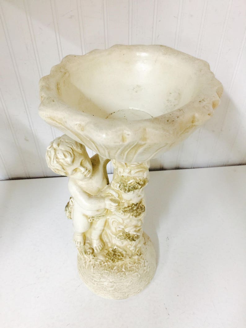 May include: Off-white cherub bird bath. The bird bath features a cherub figure holding up a scalloped bowl. The base and the tree trunk are textured with gold accents. The bird bath is made of a durable material.