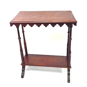 May include: A small, antique wooden side table with a rectangular top and a lower shelf. The table has decorative scalloped edges and turned legs. The wood has a warm, reddish-brown tone, and the table is set against a white background.