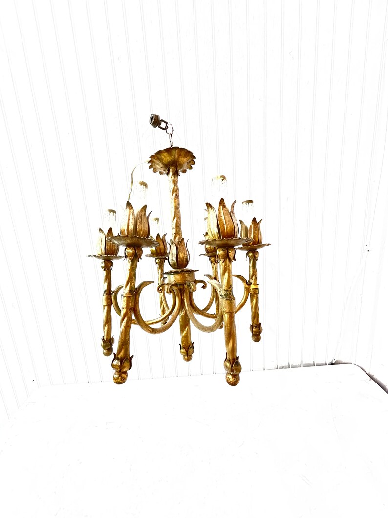 Pode incluir: Um candeeiro de teto de metal dourado com seis luzes. O candeeiro de teto tem um design floral com um caule central e seis bra&ccedil;os que se curvam para fora. Cada bra&ccedil;o tem um acess&oacute;rio de luz com uma l&acirc;mpada de vidro transparente. O candeeiro de teto est&aacute; pendurado num teto branco.