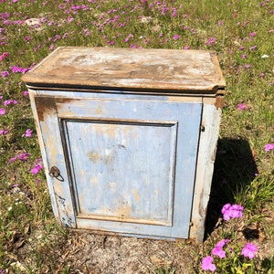 May include: A small, antique cabinet with a single door and a simple design. The cabinet is painted a light blue color and has a distressed finish. The cabinet is sitting on a bed of pink flowers.