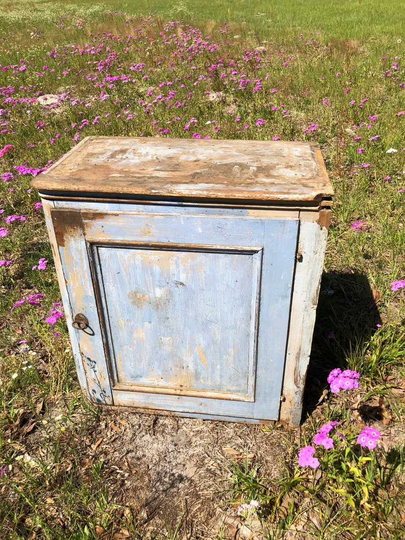 May include: A small, antique cabinet with a single door and a simple design. The cabinet is painted a light blue color and has a distressed finish. The cabinet is sitting on a bed of pink flowers.