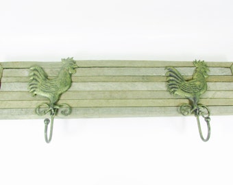 May include: A wooden wall mounted coat rack with two hooks shaped like roosters. The hooks are a green-gold color and the wood is a light brown color.