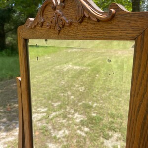 May include: A vintage wooden vanity mirror with a carved top and a beveled glass mirror. The mirror is standing on a wooden surface.