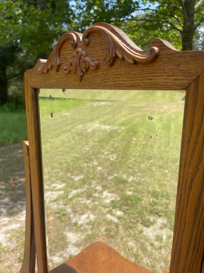 May include: A vintage wooden vanity mirror with a carved top and a beveled glass mirror. The mirror is standing on a wooden surface.
