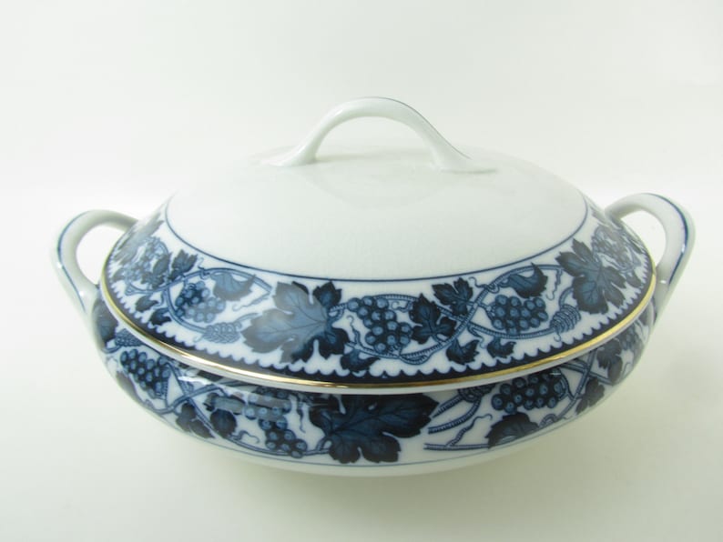 K&ouml;nnte beinhalten: Eine wei&szlig;e Porzellanservierschale mit einem blauen und goldenen Traubenranken-Design. Die Schale hat einen Deckel mit einem Griff und zwei Griffe an den Seiten.