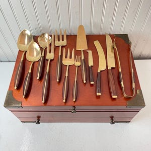 May include: A set of 14 gold-toned serving utensils with wooden handles in a wooden box. The utensils include forks, spoons, and a knife.