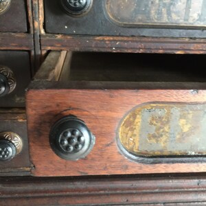 May include: A vintage wooden drawer unit with multiple drawers. Each drawer has a black knob and a label with faded text. The drawer in focus has a label that reads "F.L.T."