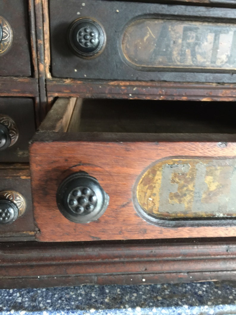May include: A vintage wooden drawer unit with multiple drawers. Each drawer has a black knob and a label with faded text. The drawer in focus has a label that reads "F.L.T."