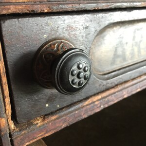 May include: Close-up of a drawer pull with a black knob and ornate brass trim. The drawer is made of dark wood with a worn finish.