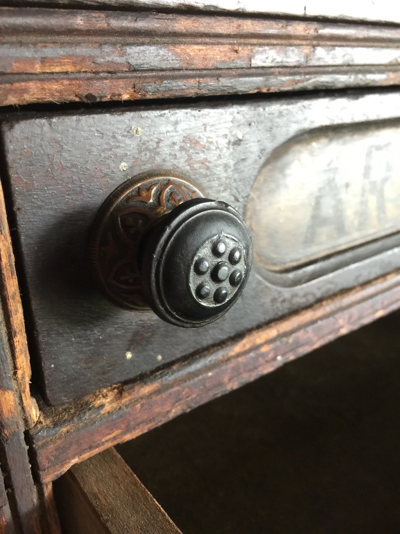 May include: Close-up of a drawer pull with a black knob and ornate brass trim. The drawer is made of dark wood with a worn finish.