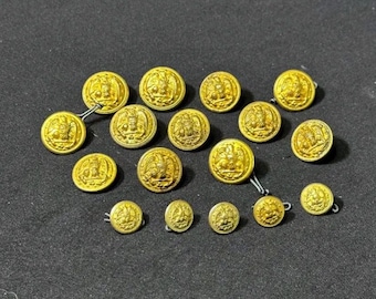 Könnte beinhalten: Eine Sammlung goldfarbener Metallknöpfe mit detaillierten geprägten Designs. Die Knöpfe sind unterschiedlich groß und weisen ein zentrales Emblem auf, das von komplizierten Mustern umgeben ist. Sie sind auf einer dunklen Oberfläche angeordnet.