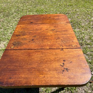 May include: A rectangular, antique wooden table with rounded corners. The table's surface is a warm, medium brown with visible wood grain and dark spots, suggesting age and use. The table is set outdoors on a bed of green grass.