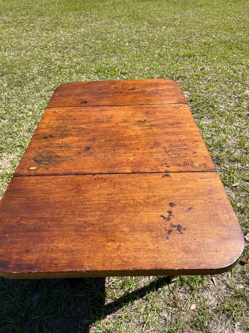May include: A rectangular, antique wooden table with rounded corners. The table's surface is a warm, medium brown with visible wood grain and dark spots, suggesting age and use. The table is set outdoors on a bed of green grass.