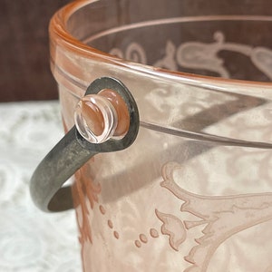 May include: A pink glass ice bucket with a silver handle and a floral design. The bucket is made of clear glass with a pink tint and has a silver metal handle.