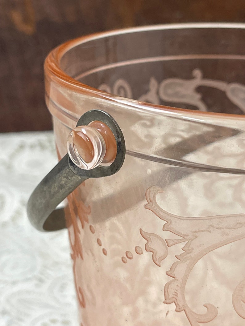 May include: A pink glass ice bucket with a silver handle and a floral design. The bucket is made of clear glass with a pink tint and has a silver metal handle.
