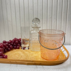 May include: A bamboo serving tray with a clear glass decanter, a clear glass tumbler, a pink glass ice bucket with a silver handle, and a bunch of red grapes.