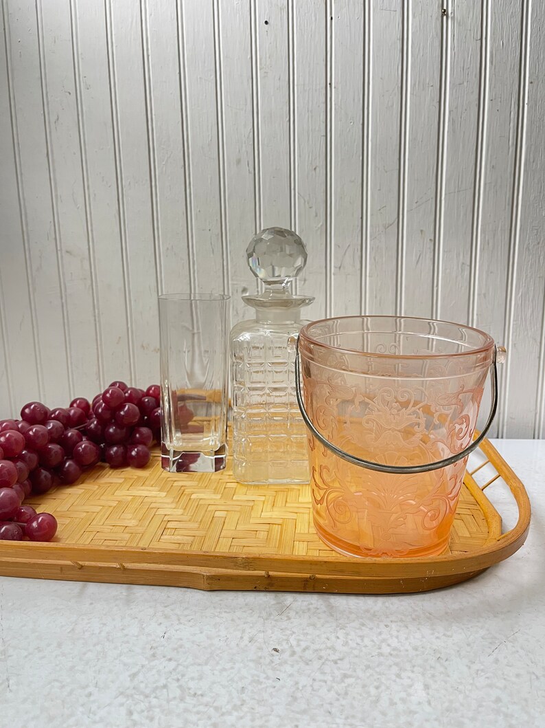 May include: A bamboo serving tray with a clear glass decanter, a clear glass tumbler, a pink glass ice bucket with a silver handle, and a bunch of red grapes.